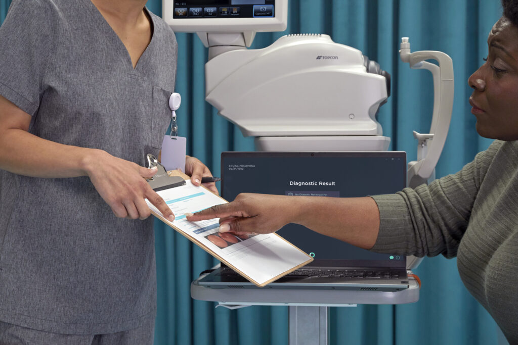 Healthcare provider and patient reviewing diagnostic results on a clipboard next to a retinal imaging device and laptop screen