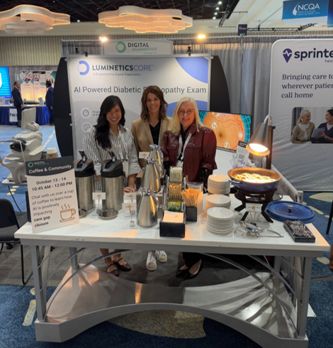 Three employees from Digital Diagnostics stand behind a coffee station at the 2025 NCQA Health Innovation Summit in front of the company’s LumineticsCore booth. A sign on the table invites attendees to “Chat with us over a cup of coffee to learn how AI is positively impacting care gap closure.”